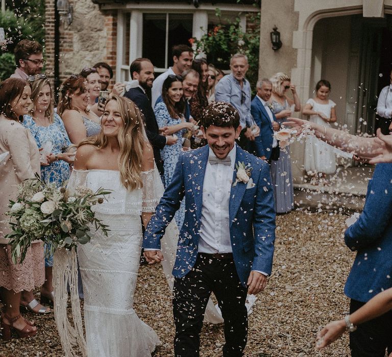 Bride in Grace Loves Lace bardot wedding dress and veil holding white and green bridal bouquet with macrame tie holds hands with groom in blue suit jacket and bow tie and guests throw confetti at Isle of Wight wedding with macrame wedding decor