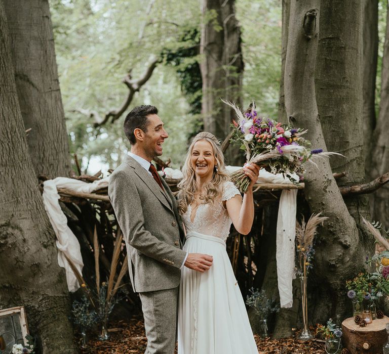 Bride in white lace cap sleeve wedding dress holding mixed bridal bouquet walks holding hands with groom in grey three piece herringbone suit and red tie in woodland at late summer wedding in Norfolk