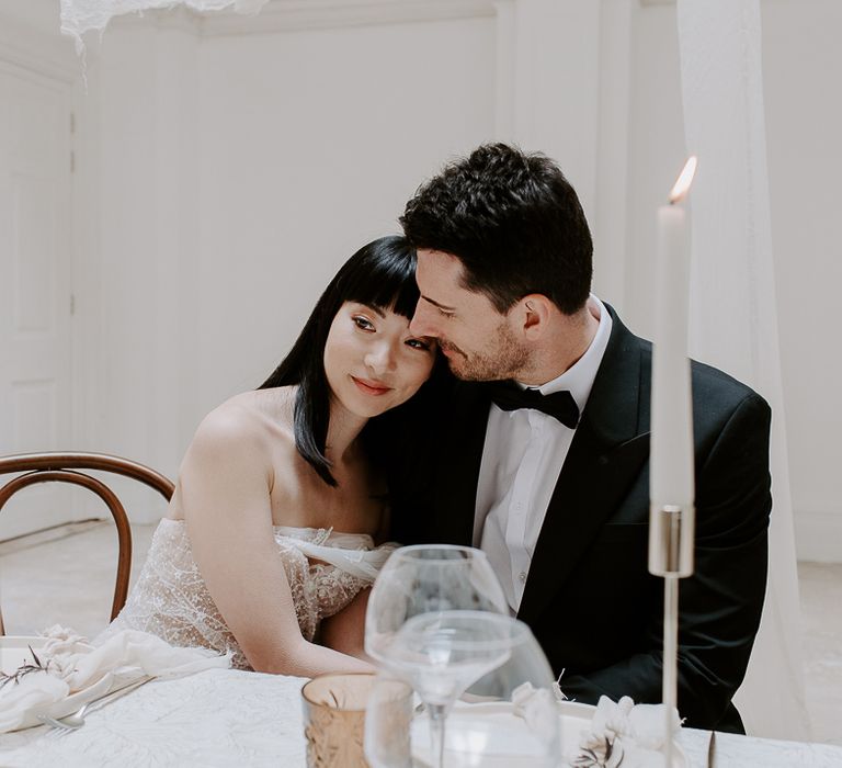 Groom in a tuxedo embracing his bride with shoulder length hair and a fringe at the reception table decorated with candles 