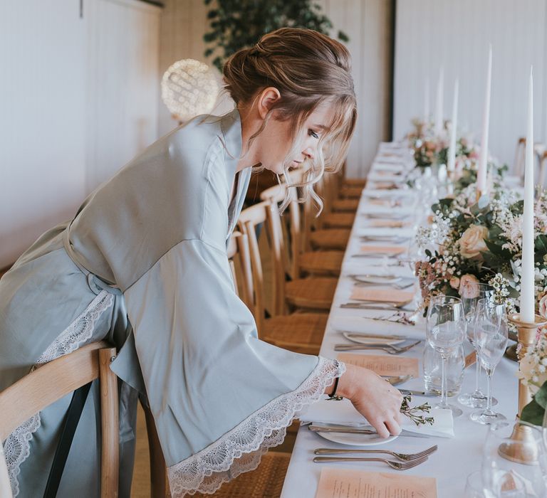 Bride in blue satin dressing gown arranges table place settings on long table in barn at Primrose Hill Farm for summer wedding