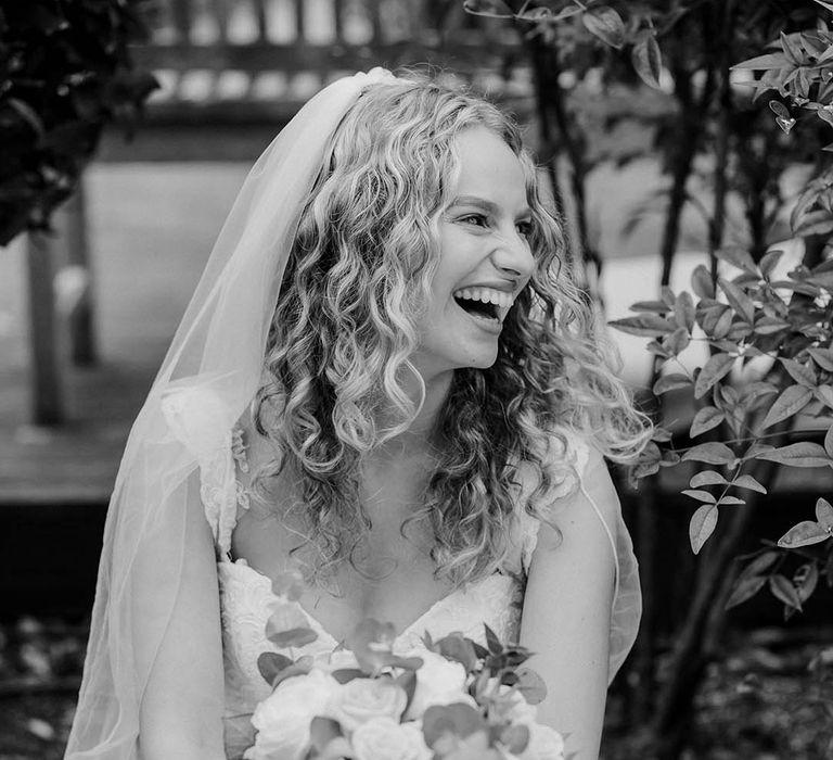 Black & white image of bride laughing with her hair in natural curls whilst holding bridal bouquet