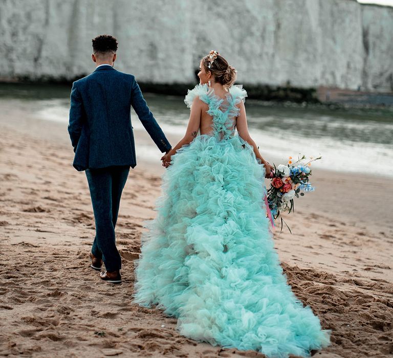 Bride in a Millia London mint green wedding dress walking along the beach with her husband in a blue wool suit 