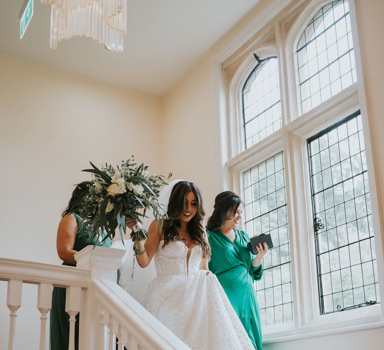 Bride in applique Pronovias wedding dress and Jimmy Choo wedding shoes walks down stairs holding green and white rose wedding bouquet with bridesmaids in green bridesmaid dresses at Notley Abbey before her wedding ceremony