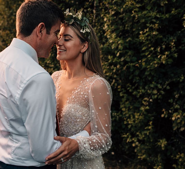 Bride in long mesh sleeve wedding dress with v neck and flower crown smiles whilst holding groom in white shirt and red tie during golden hour