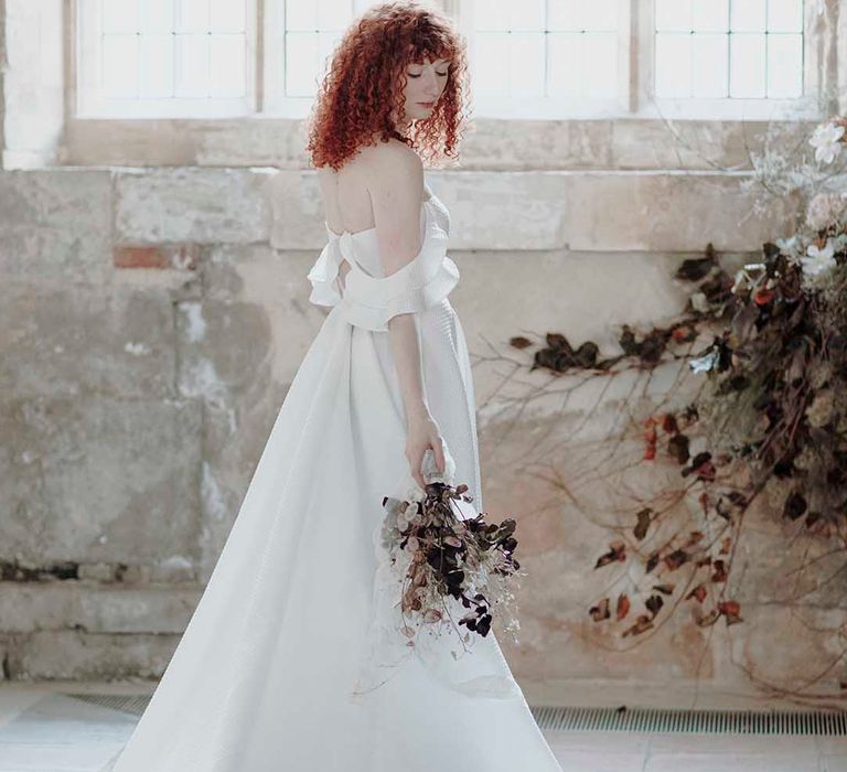 Bride with naturally curly red hair standing in Blackfriars Priory in a cold shoulder wedding dress holding a dried flower bouquet 