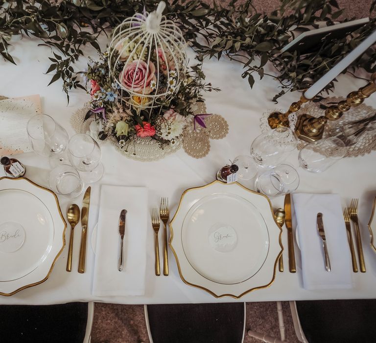 Rustic chic wedding reception with gold rimmed tableware, foliage garland, bright florals in a cage and candlesticks 