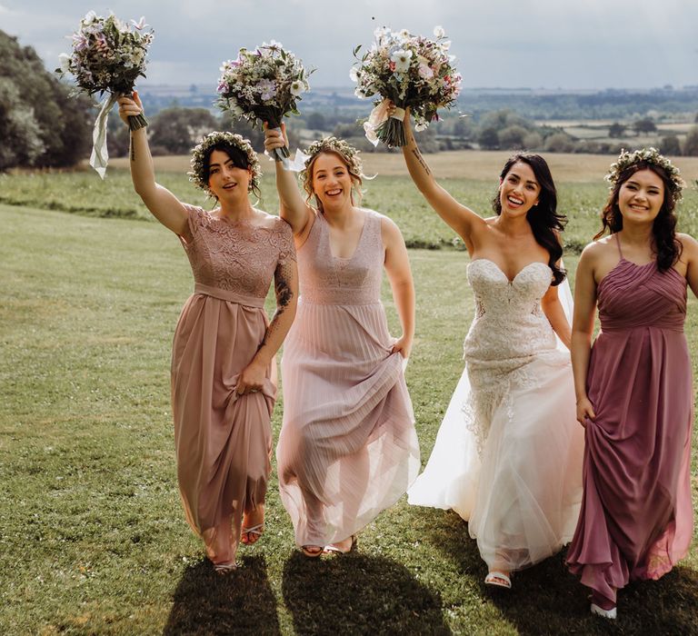 Bride in lace and tulle wedding dress with sweetheart neckline walks with bridesmaids in mixed pink bridesmaid dresses through field whilst they hold up multicoloured pastel wedding bouquets