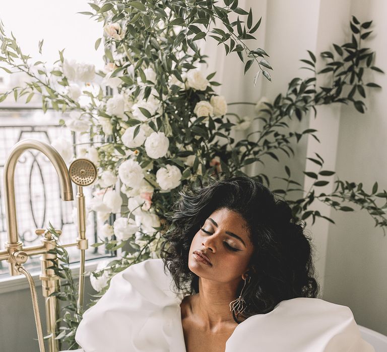 Beautiful bride in a puff sleeve wedding dress with plunging neckline sitting in a bath surrounded bu white flowers and foliage 