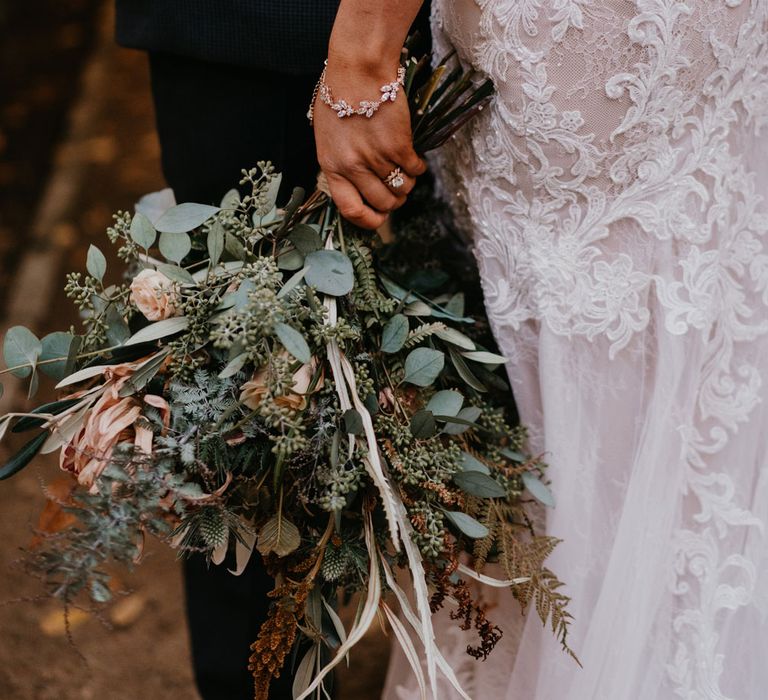 Bride in lace and tulle wedding dress wearing silver bracelet and large mixed bridal bouquet stands with groom in dark suit 