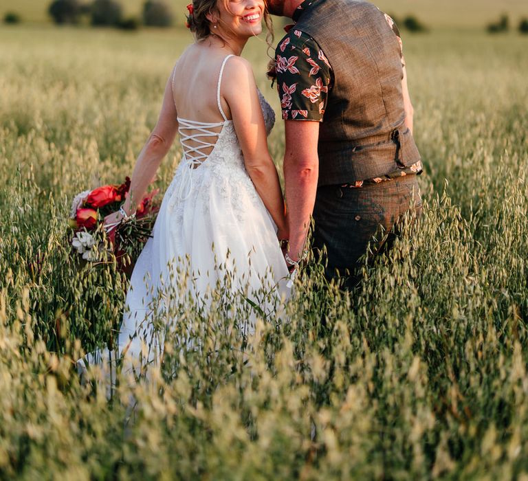 Bride in corset back wedding dress holding rustic bouquet smiles as she stands in long grass field with groom in patterned short sleeve shirt and waistcoat