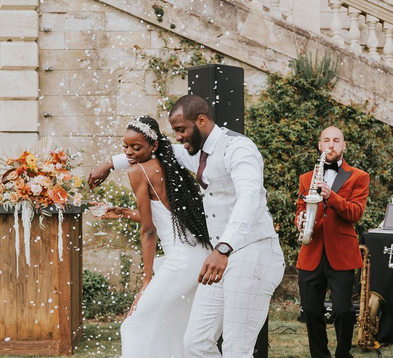 Black bride in a beaded wedding dress and groom in a stone check suit facing outside at Kirtlington Park with confetti falling all around them 