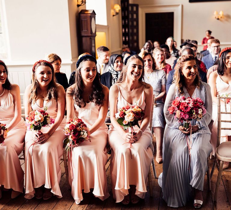 Bridesmaids in baby pink satin bridesmaid dress, custom dark pink headbands and pink, red and white bouquets sit in the front row of Elmore Court wedding ceremony