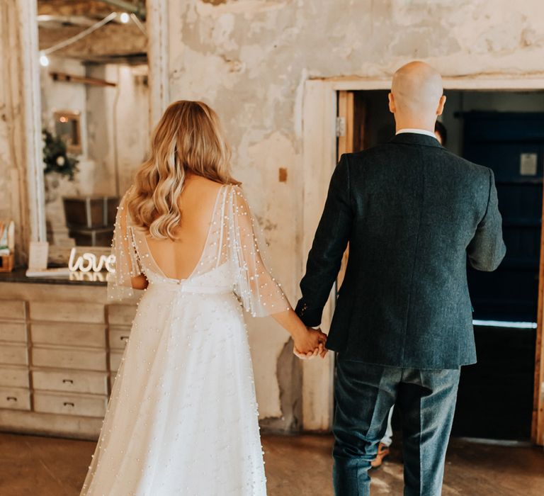 Bride in love v back wedding dress with pearls and train walks out of ceremony holding hands with groom in tweed suit and brown shoes