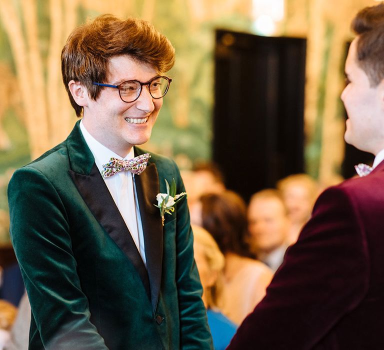 Smiling groom in forest green velvet tuxedo jacket and floral bow tie with rounded glasses 