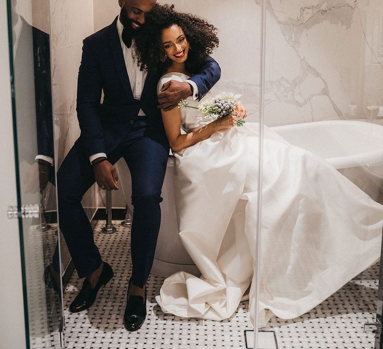 Black groom in a navy blue suit and bow tie embracing his Black bride in a one shoulder wedding dress in the bathroom at Mayfair Townhouse 