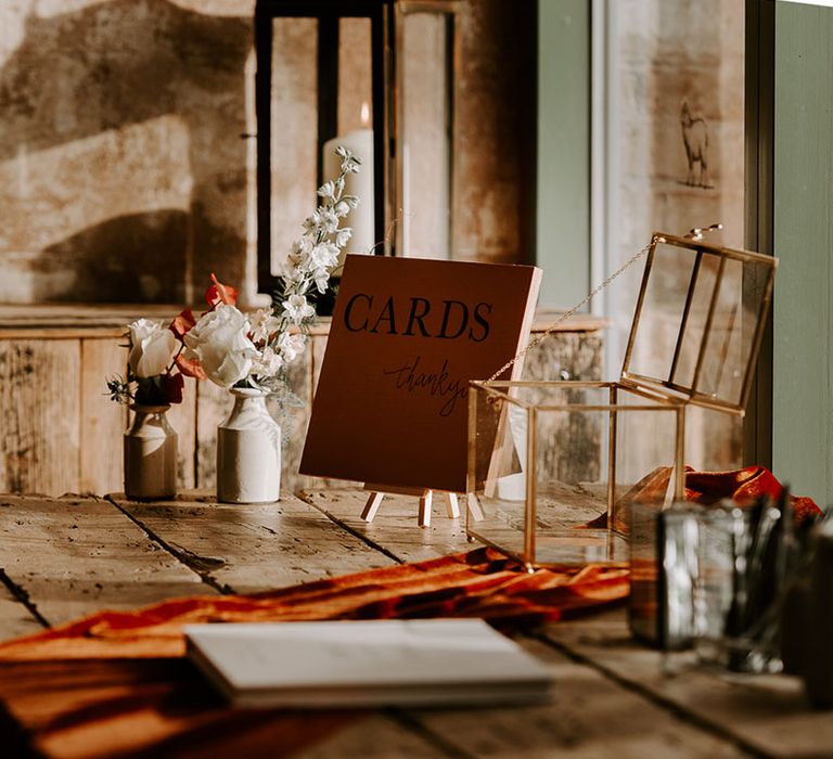 Wedding guestbook and card signing station with pot of pens and orange sign with gold rimmed glass box 