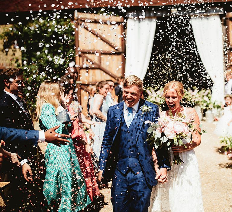 Bride holding pink rose bouquet and hands with the groom exit their ceremony to white confetti