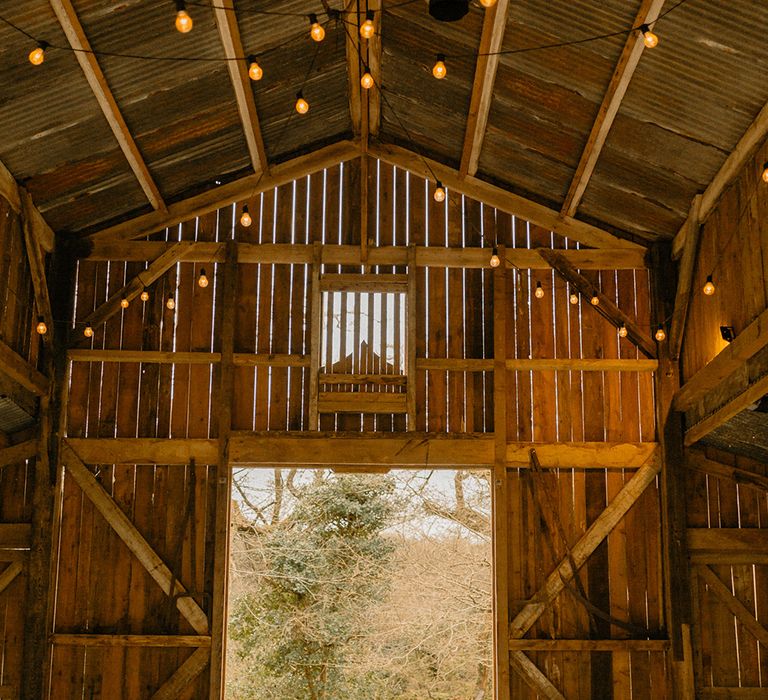 Silver lanterns with candles and hanging naked lightbulbs with vintage milk churns full of autumnal orange flowers for rustic barn wedding