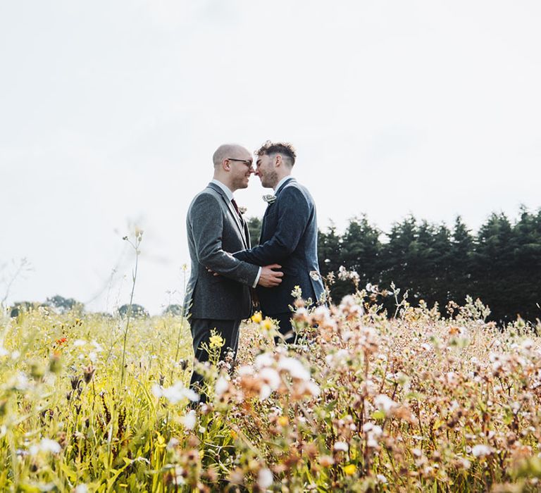 Grooms lean in for a kiss together as they stand in a field in grey and blue suit jackets