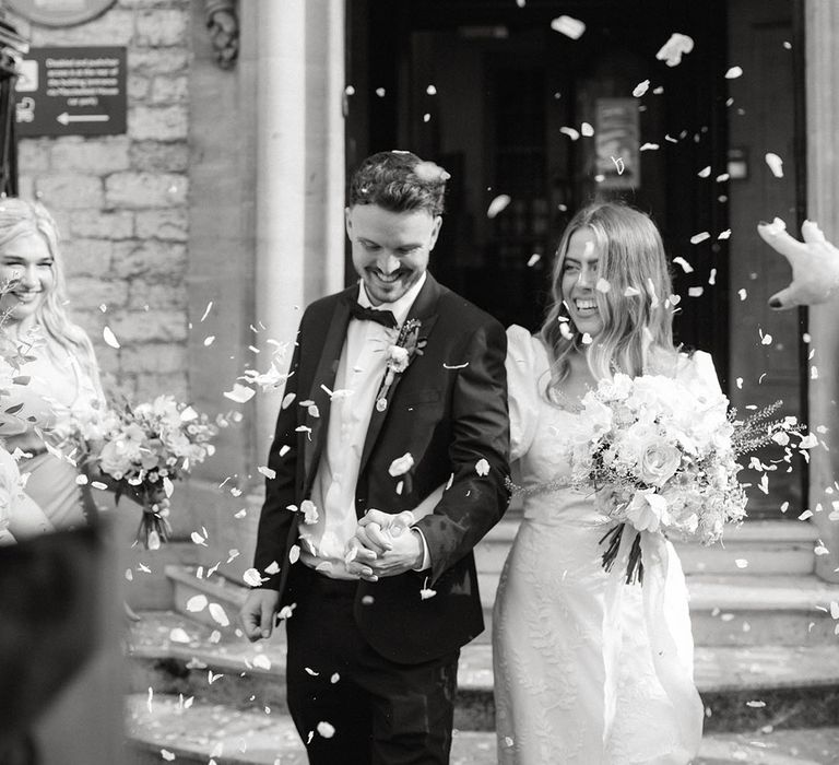 Bride in patterned puff sleeve wedding dress has confetti exit with groom in black tie holding colourful bouquet 