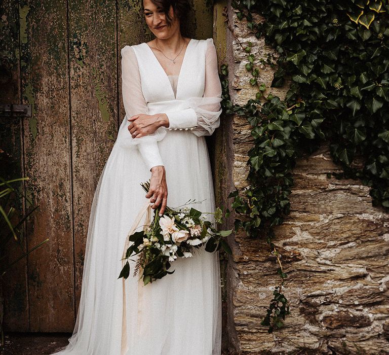 Bride leans against a wall wearing sheer long sleeve wedding dress by Sassi Holford