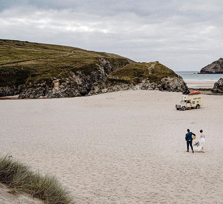 Bride and groom walk along Cornwall beach together with van in distance