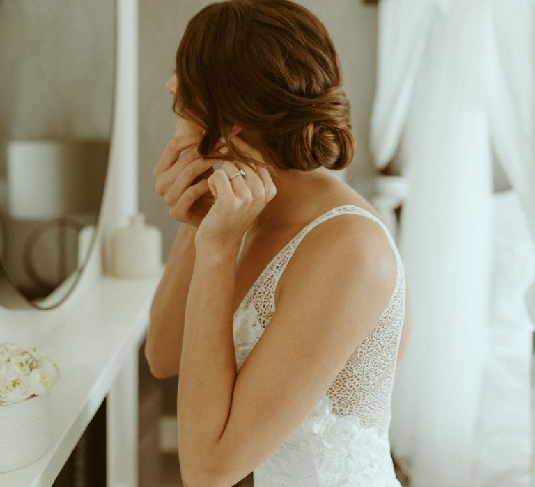 Bride in lace wedding dress and brown hair in an updo puts in her earrings for her wedding day