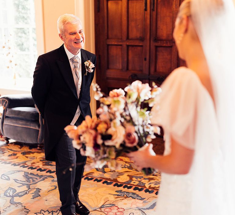 Father of the bride in three piece morning suit with light tie smiles as he sees the bride for the first time 