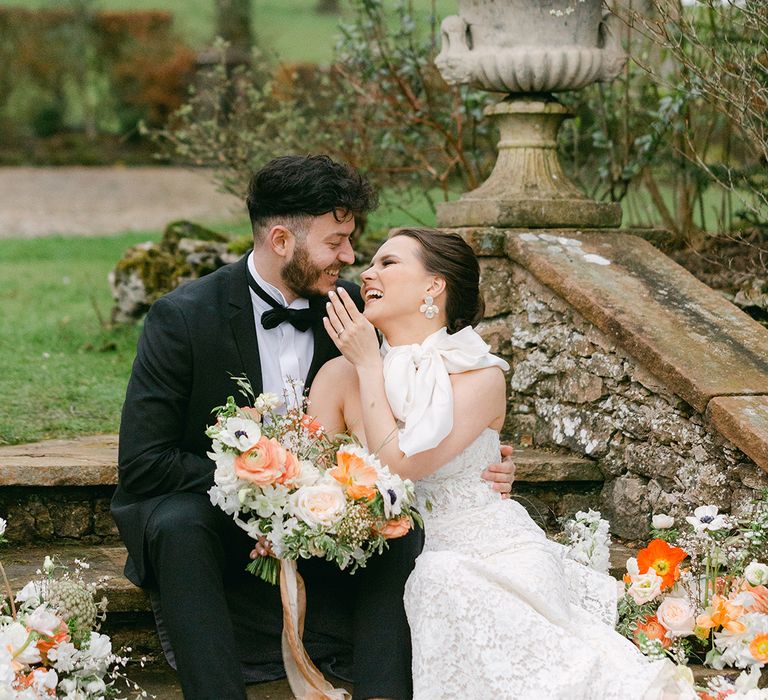 Bride in a lace wedding dress sitting on the steps at Holesfoot with her groom in a tuxedo surrounded by peach and white flowers 