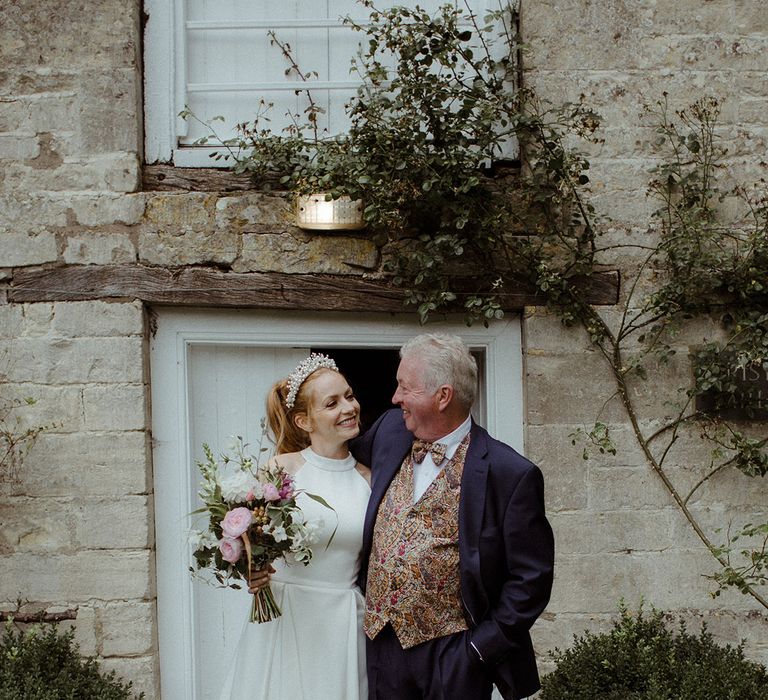 Bride in pleated wedding dress with pearl headband stands with groom in paisley patterned waistcoat and tie 