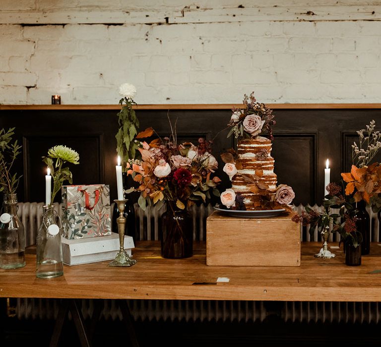 Rustic naked wedding cake sits on wooden table surrounded by florals 