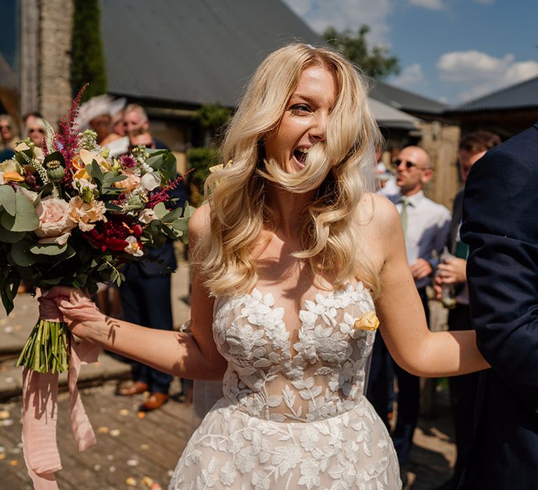 Bride with softly curled blonde hair in strapless lace Lillian West wedding dress walks through confetti with groom in blue suit 