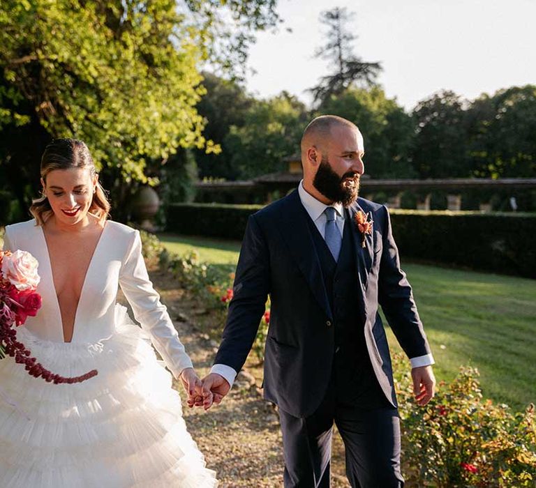 Bride & groom hold hands outdoors on their wedding day as bride holds bright floral bouquet matching her husbands buttonhole 