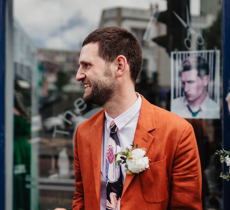 Groom wears orange Paul Smith linen suit complete with white shirt, floral illustrated tie and white floral buttonhole 