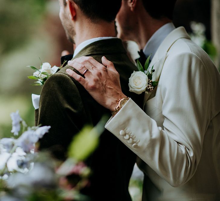 Groom wearing white black tie rests his hand on his groom who wears green velvet jacket, both wearing white rose floral buttonholes