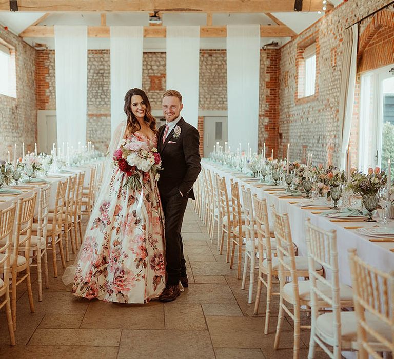 Bride in flower wedding dress and groom in a brown suit smile at their wedding reception with pink taper candles, green glassware and copper cutlery