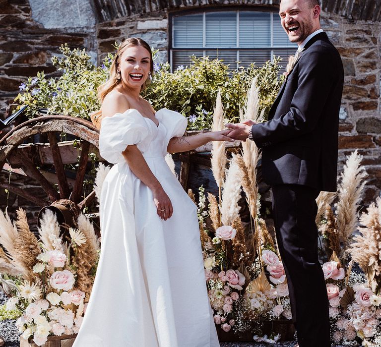 Groom smiles as puts on the bride's wedding ring who wear a puffy off the shoulder wedding dress with pampas grass and pink flowers decor 