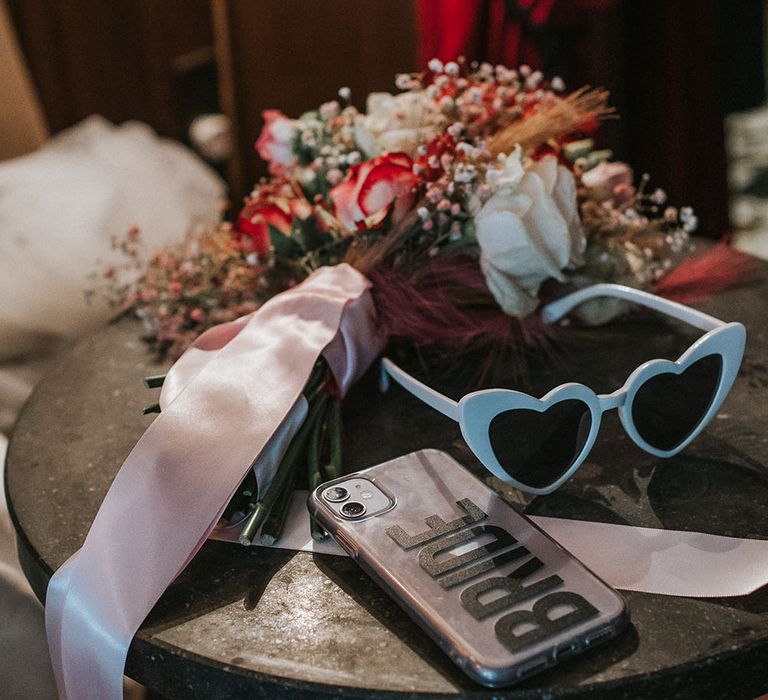 Heart-shaped sunglasses beside bright floral bouquet tied with pink ribbon 