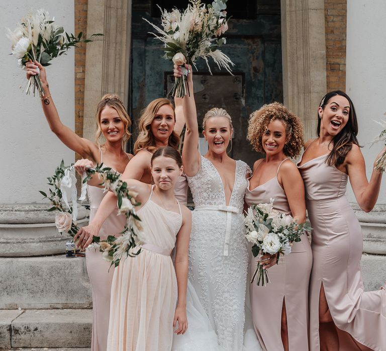 Bride stands with bridesmaids and flower girl in pastel pink dresses holding white floral bouquets with pampas grass 