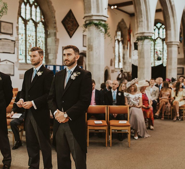 Groom stands in a grey and blue morning suit with his groomsmen at the altar for the church wedding ceremony