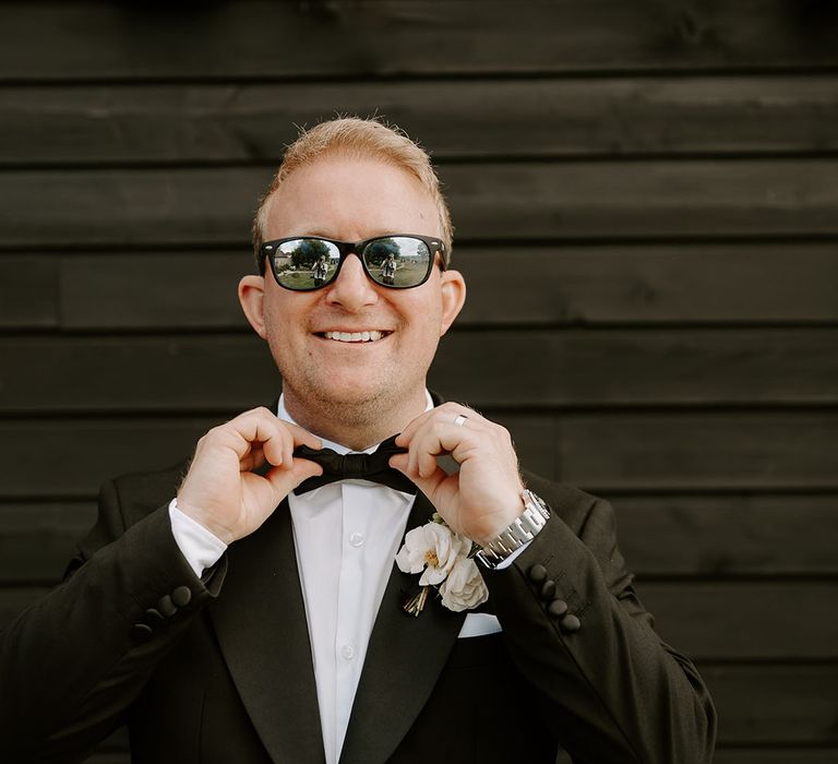 Groom wears black-tie with sunglasses and white floral buttonhole 