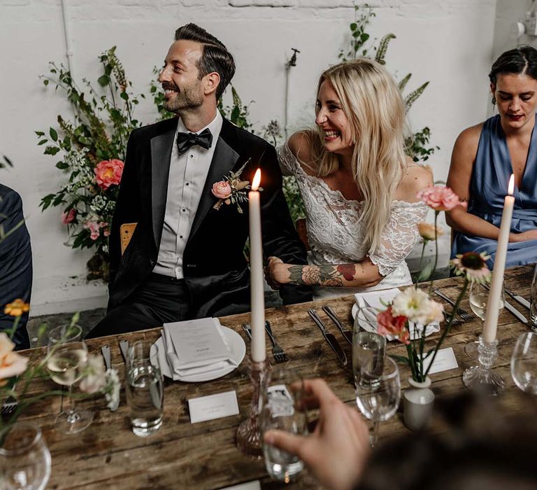 Bride in lace wedding top and groom in dark tux and bowtie laughing at wedding speeches in modern stylish reception room