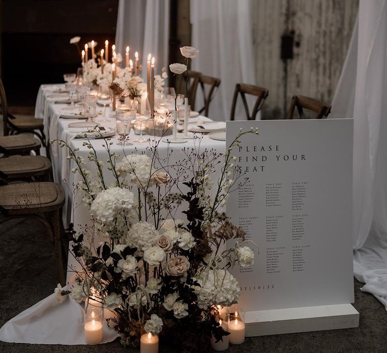 Neutral minimalistic table plan and wedding tablescape with artsy neutral toned floral decorations, long tapered beige candles, light grey ceramic plates and candle holders and a white cotton table runner