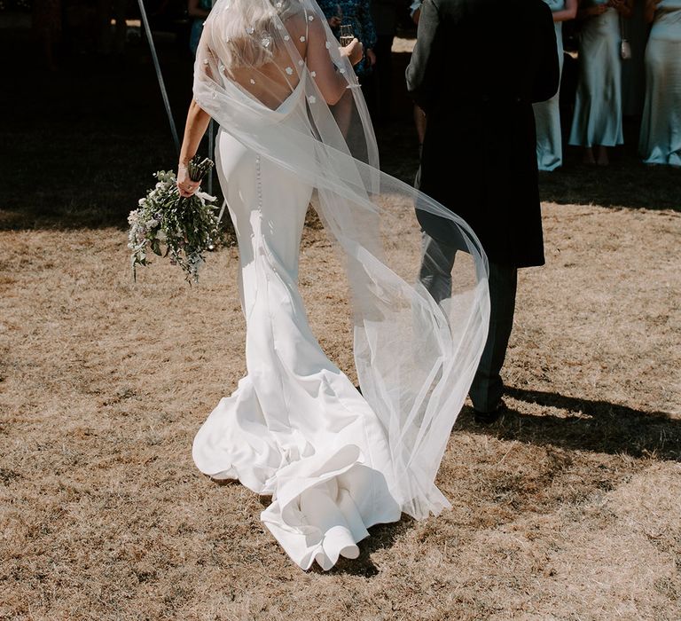 Bride in cowl back wedding dress with flower applique veil walking with the groom to the marquee reception 