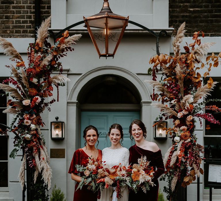 Bride in long sleeve lace vintage wedding dress and closed toe satin heels standing with bridesmaids in mismatched velvet bridesmaid dresses holding large autumnal bouquets standing under large floral arch with roses, peonies, foliage and pampas grass