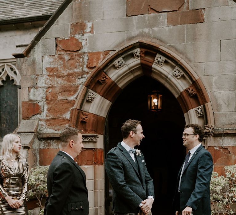 Groom and groomsmen stand outside the church as the guests arrive wearing kilts for Scottish traditional church wedding ceremony 