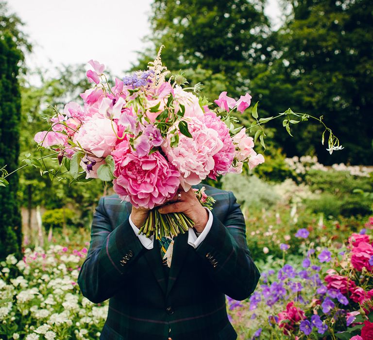 Groom dark forest green tartan suit with patterned tie, white pocket square and pink and green boutonniere holding up pink floral bouquet with pink peonies, fuchsia roses, lilac sweet peas and green foliage