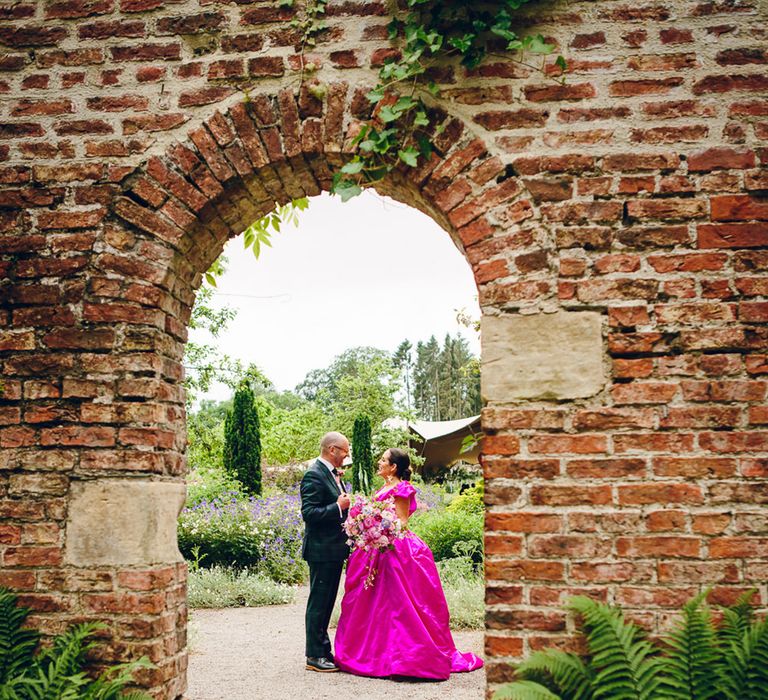 Bride in off the shoulder front ruching pink wedding dress smiling and talking to groom in in dark forest green tartan suit with patterned tie, white pocket square and pink and green boutonniere through an exposed brick archway in Middleton Lodge