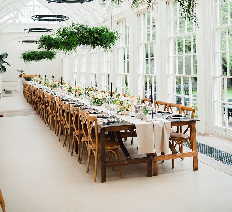 Light and bright reception room with neutral off-white table runners, rose and foliage floral arrangements, pink and black tapered candles, black candlestick holders and suspended foliage decorations