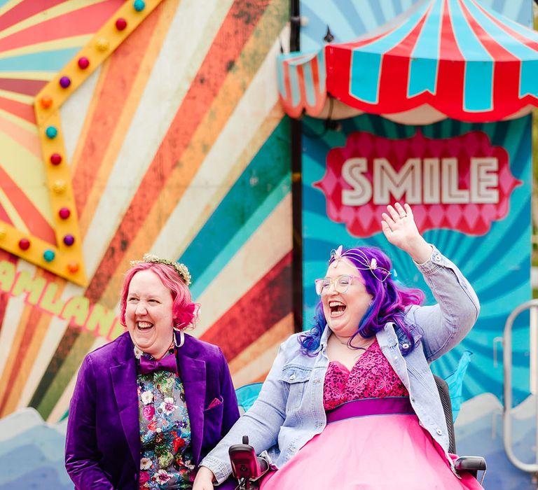 Bride in wheelchair wearing personalised denim jacket, pink wedding dress and blue tights holding hands with bride in purple velvet blazer and black suit trousers with purple bowtie, pink patterned pocket square and colourful patterned shirt